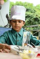 linda indio cocinero chico preparando helado con frutas y nueces plato como un parte de no fuego Cocinando cuales incluye vainilla hielo crema, duende, Coco polvo, recién Cortado frutas y fresa jarabe. pequeño niño preparando comida foto