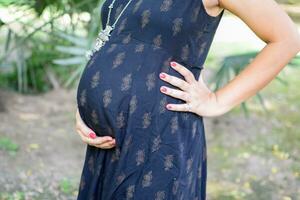 un embarazada indio dama poses para al aire libre el embarazo disparar y manos en barriga, indio embarazada mujer pone su mano en su estómago con un maternidad vestir a sociedad parque, embarazada fuera de maternidad disparar foto