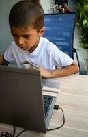 pequeño chico sentado a mesa utilizando ordenador portátil para en línea clase en grado 1, niño estudiando en ordenador portátil desde hogar para distancia aprendizaje en línea educación, colegio chico niños estilo de vida concepto foto