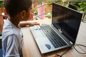 pequeño chico sentado a mesa utilizando ordenador portátil para en línea clase en grado 1, niño estudiando en ordenador portátil desde hogar para distancia aprendizaje en línea educación, colegio chico niños estilo de vida concepto foto