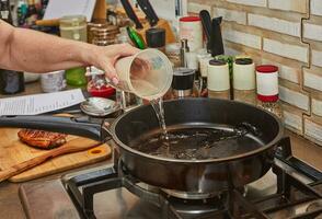 Chef pours water from measuring cup into frying pan on gas stove photo