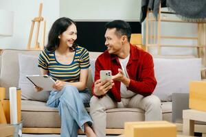 asiático joven atractivo Pareja hombre y mujer utilizar tableta y teléfono inteligente en línea compras mueble Decorar casa con caja de cartón paquete moverse en nuevo casa. joven casado Moviente hogar. foto