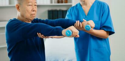 asiático fisioterapeuta Ayudar mayor hombre paciente extensión brazo durante ejercicio correcto con pesa en mano durante formación mano con paciente espalda problemas foto