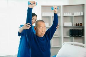 asiático fisioterapeuta Ayudar mayor hombre paciente extensión brazo durante ejercicio correcto con pesa en mano durante formación mano con paciente espalda problemas foto