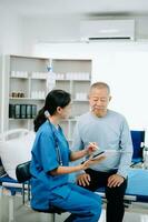 Asian doctor in white suit take notes while discussing and Asian elderly, man patient who lying on bed with receiving saline solution in hospital or clinic. photo