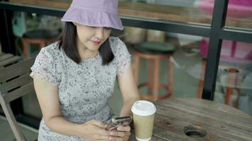 Frau Typen ein Botschaft auf ein Smartphone im ein Kaffee Geschäft und sie sitzt beim ein Tabelle mit Kaffee mit ein Handy, Mobiltelefon Telefon. video