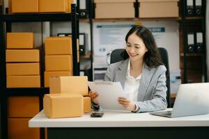 Startup small business SME, Entrepreneur owner woman using smartphone or tablet taking receive and checking online purchase shopping order to preparing pack product box. photo