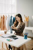 Successful fashion designer. Attractive young asian woman with smile while standing in workshop. Beautiful owner business woman working and holds tablet, laptop and smartphone on desk in studio. photo