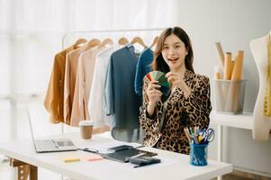 Successful fashion designer. Attractive young asian woman with smile while standing in workshop. Beautiful owner business woman working and holds tablet, laptop and smartphone on desk in studio. photo