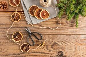 Homemade garland of dried oranges on a wooden rustic table, vintage scissors, a skein of thread for crafts, spruce branches. Top view. photo