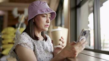 de edad mediana mujer participación un café jarra y mirando a un teléfono inteligente mientras sentado video