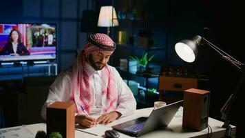 Muslim man at home paying attention in elearning seminar teleconference with teacher, writing on notepad. Middle Eastern homeschooling student listening to lesson while doing his homework tasks video