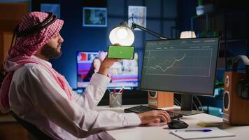 Arab man watching video on green screen cellphone while looking at business annual report charts on computer. Worker spending time on chroma key mobile phone while solving job tasks at home