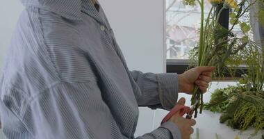 vrouw aan het doen nieuw boeket. meisje werken in bloem winkel studio. vrouw bloemist artiest reinigt bloemen voor een mooi boeket, werkplaats visie. video