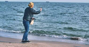 Blonde girl in jeans on the shore of the Black Sea throws pebbles into the sea against the backdrop of ships in the roadstead video