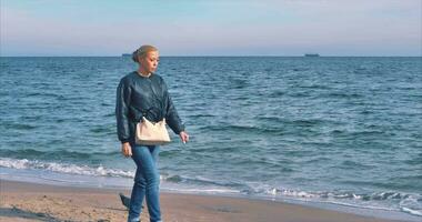 Against the backdrop of the Black Sea coast, a girl in jeans with a white bag walks along the sand video