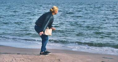 un' bionda ragazza nel jeans con un' bianca Borsa passeggiate lungo il riva di il nero mare e lanci ciottoli in il mare video