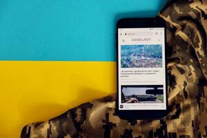 KYIV, UKRAINE - 4 MAY, 2023 Zaxid Net ukrainian news portal on smartphone screen with ukrainian flag and camouflage fabric photo