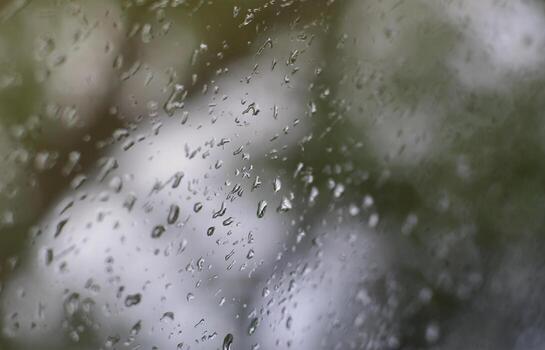 A photo of rain drops on the window glass with a blurred view of the blossoming green trees. Abstract image showing cloudy and rainy weather conditions