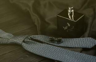 A bottle of mens cologne and cufflinks with blue tie lie on a black luxury fabric background on a wooden table. Mens accessories photo