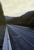 Road in autumn forest at sunset in Carpathian mountains, Ukraine. Beautiful mountain roadway with orange tress and high rocks. Landscape with empty highway through the woods in fall photo