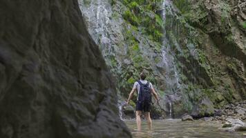 Man with backpack spreading hands in front of waterfall. video