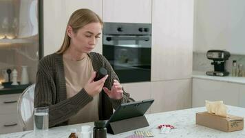 A woman reads instructions for using pills using a gadget while sitting at a table in the kitchen. video