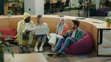 multiétnico negocio equipo de compañeros de trabajo que se discute un proyecto mientras sentado en otomanos en un moderno oficina.diferente razas diversas gente creativa equipo,negocio socios video