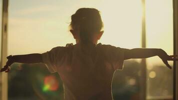 The child holds his hands to the sides while standing near the window during dawn. video