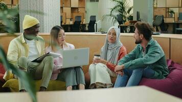 Multi-éthnique affaires équipe de collègues de travail discuter une projet tandis que séance sur poufs dans une moderne bureau.différent courses, diverses les gens, créatifs équipe, entreprise les partenaires video