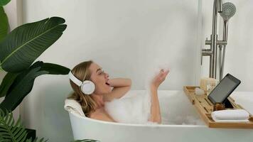 uma mulher vestindo fones de ouvido escuta para música enquanto deitado dentro a banheira durante água procedimentos. video