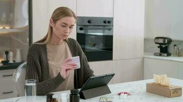 uma mulher lê instruções para usando pílulas usando uma gadget enquanto sentado às uma mesa dentro a cozinha. video