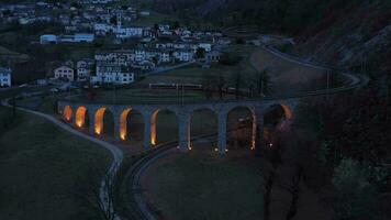 Train on Illuminated Brusio Spiral Viaduct in Switzerland in the Evening. Bernina Railway. Swiss Alps. Aerial View. Drone Flies Backwards and Upwards video