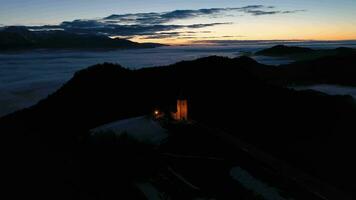 Illuminated Church of St. Primoz and Felicijan at Dawn above the Clouds. Julian Alps. Jamnik, Slovenia, Europe. Aerial View. Orbiting video