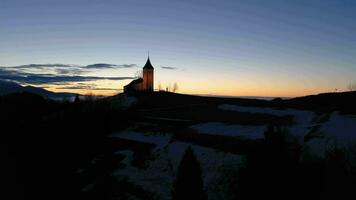 Illuminated Church of St. Primoz and Felicijan at Dawn above the Clouds. Julian Alps. Jamnik, Slovenia, Europe. Aerial View. Drone Flies Forward and Upwards at Low Level video