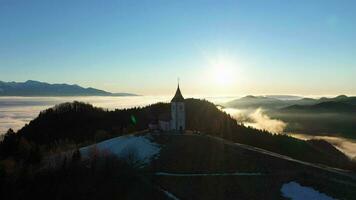 Church of St. Primoz and Felicijan at Sunrise above the Clouds. Julian Alps. Jamnik, Slovenia, Europe. Aerial View. Orbiting video