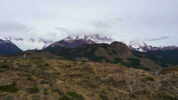 homem com laranja mochila em pé e olhando às montar fitz Roy dentro nuvens. el Chalten cidade. colinas e coberto de neve montanhas. andes, Patagônia, Argentina. aéreo visualizar. zangão moscas frente às baixo nível video