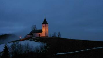 verlichte kerk van st. primo en felicijan Bij winter avond mistig schemering. bewolkt lucht. Jamnik, Slovenië. antenne visie. in een baan om de aarde video