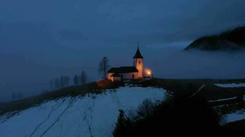 illuminé église de st. primez et Félicijan à hiver soir brumeux crépuscule. jamnik, Slovénie. aérien voir. en orbite video