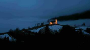 Illuminated Church of St. Primoz and Felicijan at Cloudy Winter Evening Twilight. Cloudy Sky. Jamnik, Slovenia, Europe. Aerial View. Drone Flies Forward video