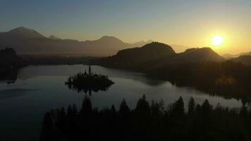 desangrado lago y marijinega vnebovzetja Iglesia a amanecer. dorado hora. colinas y bosque. Julian Alpes. Eslovenia, Europa. reflexión en lago. aéreo vista. zumbido moscas oblicuo video