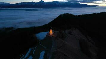 Illuminated Church of St. Primoz and Felicijan in the Morning Twilight above the Clouds. Julian Alps. Jamnik, Slovenia, Europe. Aerial View. Orbiting video