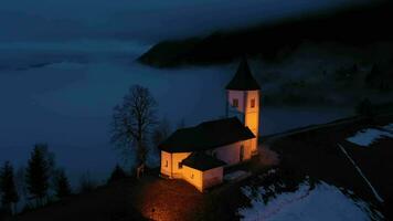 verlichte kerk van st. primo en felicijan Bij bewolkt winter nacht. bewolkt lucht. Jamnik, Slovenië, Europa. antenne visie. dar vliegt achteruit en omhoog video