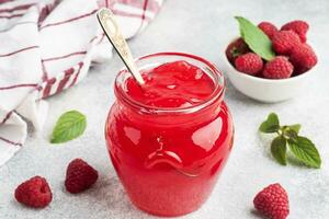 Homemade raspberry jam in a glass jar and fresh raspberries with mint on a grey concrete background. Copy space photo