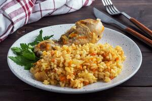 Delicious Asian pilaf, stewed rice with vegetables and chicken drumstick on a plate. Wooden rustic background. photo