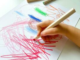 small child draws with colored pencils on paper on white table. photo