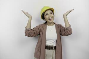 Excited Asian woman employee wearing safety helmet pointing at the copy space above her, isolated by white background photo