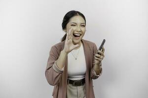 A young beautiful Asian woman employee wearing cardigan and holding her phone is shouting and screaming loud with a hand on her mouth, isolated by white background. photo