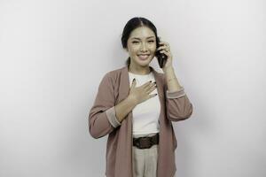 contento consciente de agradecido joven asiático mujer empleado vistiendo cárdigan y teniendo un teléfono llamada y mano en cofre sonriente aislado en blanco antecedentes sensación No estrés, foto