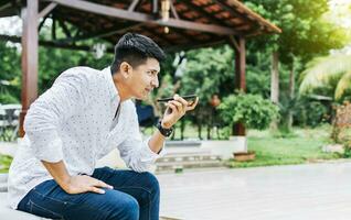 Handsome man sitting outside sending a voice message with his cell phone, Teenage sitting calling on the phone outside, Attractive man sitting sending a voice message with his phone and gesturing photo